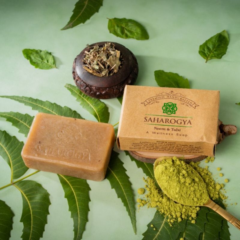 A bar of Saharogya Neem & Tulsi soap, its packaging, a wooden bowl with dried leaves, green moringa powder on a metal spoon, and fresh neem leaves on a light green background. The soap is rectangular with smooth edges and an earthy color. The box displays the product name in sans-serif type. All items are arranged flatly for an overhead shot; no people or hands appear.