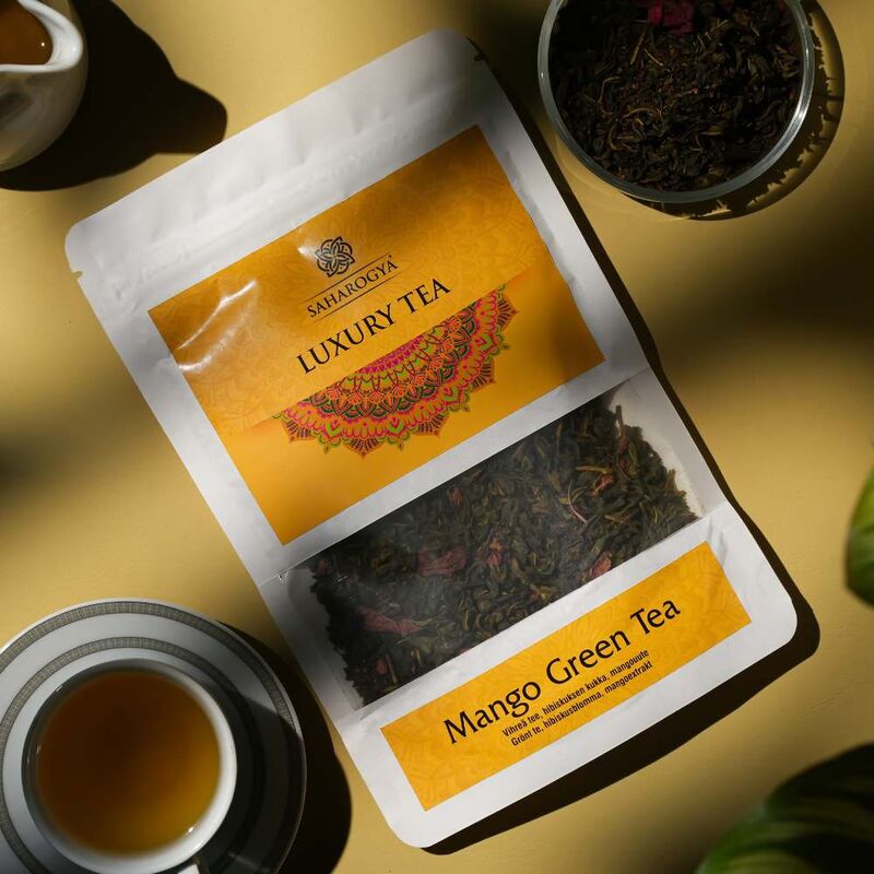 Saharogya Mango Green Tea; valko-keltainen pussi merkitty "Luxury Tea" ja "Mango Green Tea" pöydällä valkoisen teekupin ja pyöreän valkoisen kulhon vieressä, kulhossa irtonaista mango-vihreää teetä; pussi on suorakulmainen mattapintainen, lohkokirjaimin painettu; esineet ovat tavallisella pöytäpinnalla voimakkaassa lämpimässä auringonvalossa terävin varjoin; ei ihmisiä tai toimintaa kuvassa.