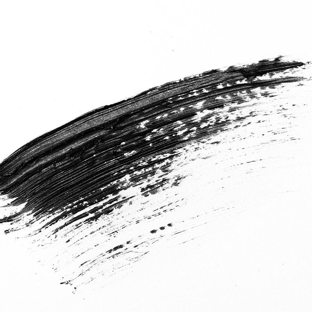 Sante Mademoiselle Sensitive Mascara; rohkea musta siveltimenveto kaartuu vinosti valkoista taustaa vasten, häipyen vähitellen ja hajoten ilmeikkäisiin roiskeisiin ja juoviin oikealla puolella; kuvassa ei erotu tuotetta, materiaalia, pintakäsittelyä, pakkausta tai etikettitekstiä; kuva on eristetty näkymä ilman ihmisiä tai käsiä.