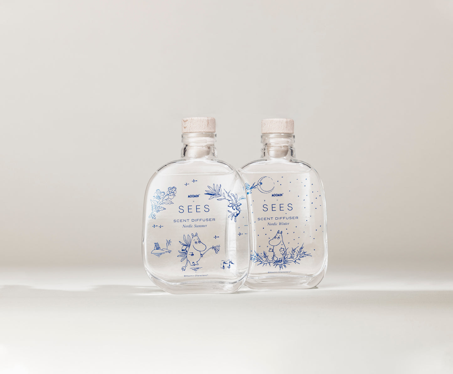 SEES x Moomin Room Diffuser Nordic Winter; kaksi kirkasta lasipulloa korkkitulpin, tekstillä SEES Scent Diffuser; kummassakin pullossa sinisiä kukka- ja pieneläinkuvituksia pinnalla; pullot läpinäkyviä ja lieriömäisiä, mattapaperiset etiketit groteskifontilla; tausta yhtenäinen valkoinen ilman muita esineitä tai tekstuureja; ei ihmisiä tai käsiä, suoraviivainen tuotekuva.