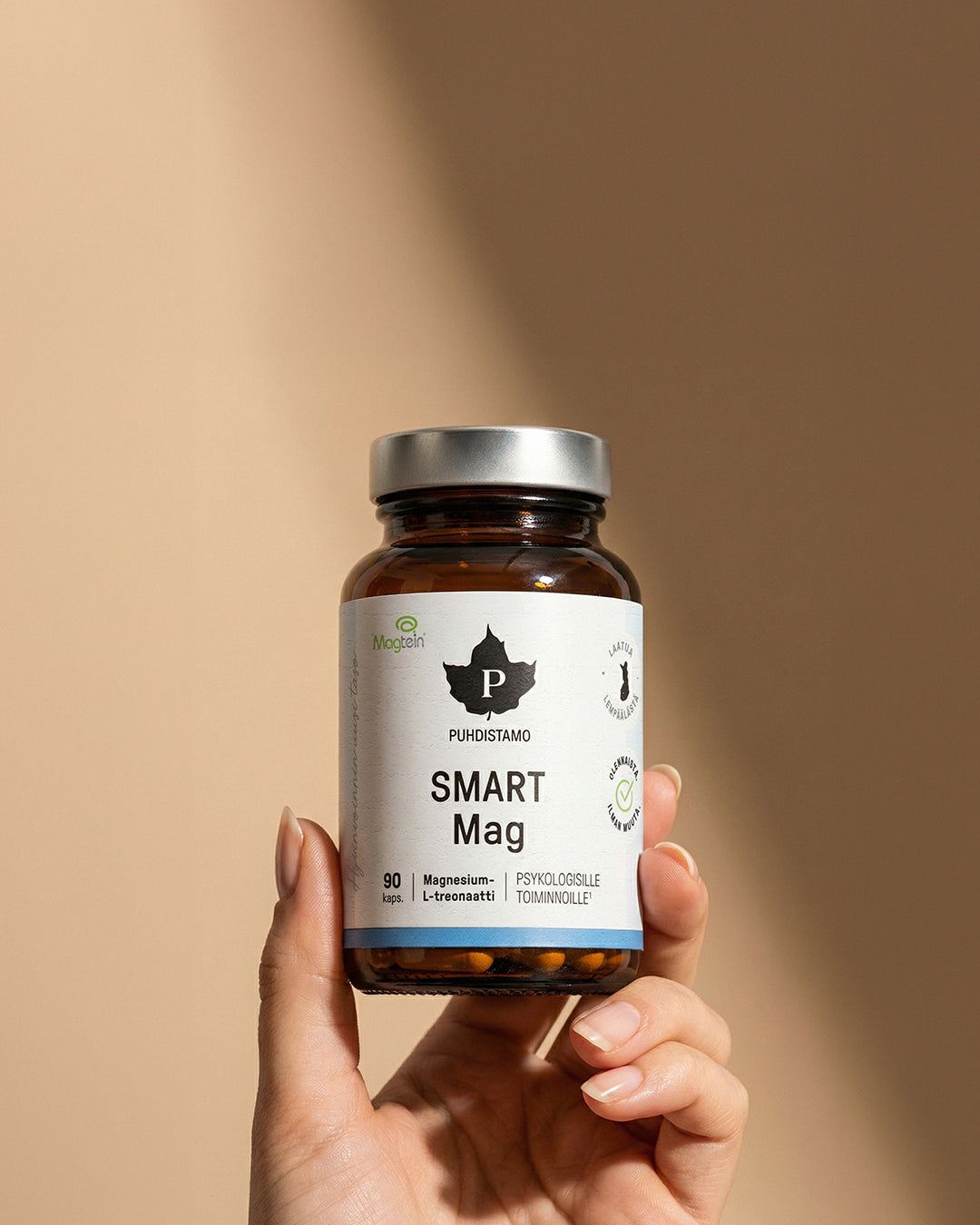 Puhdistamo Smart Mag; käsi pitelee sylinterin muotoista ruskeaa lasipurkkia sileällä mattapinnalla ja mustalla kierrekorkilla; etuetiketissä lihavoitu mustavalkoinen groteskiteksti kertoo selkeästi Puhdistamo Smart Mag plus magnesium, L-glysinaatti ja 90 vegaanista kapselia; tausta yhtenäisen beige; kuva rajattu tiukasti käden ja purkin ympärille.