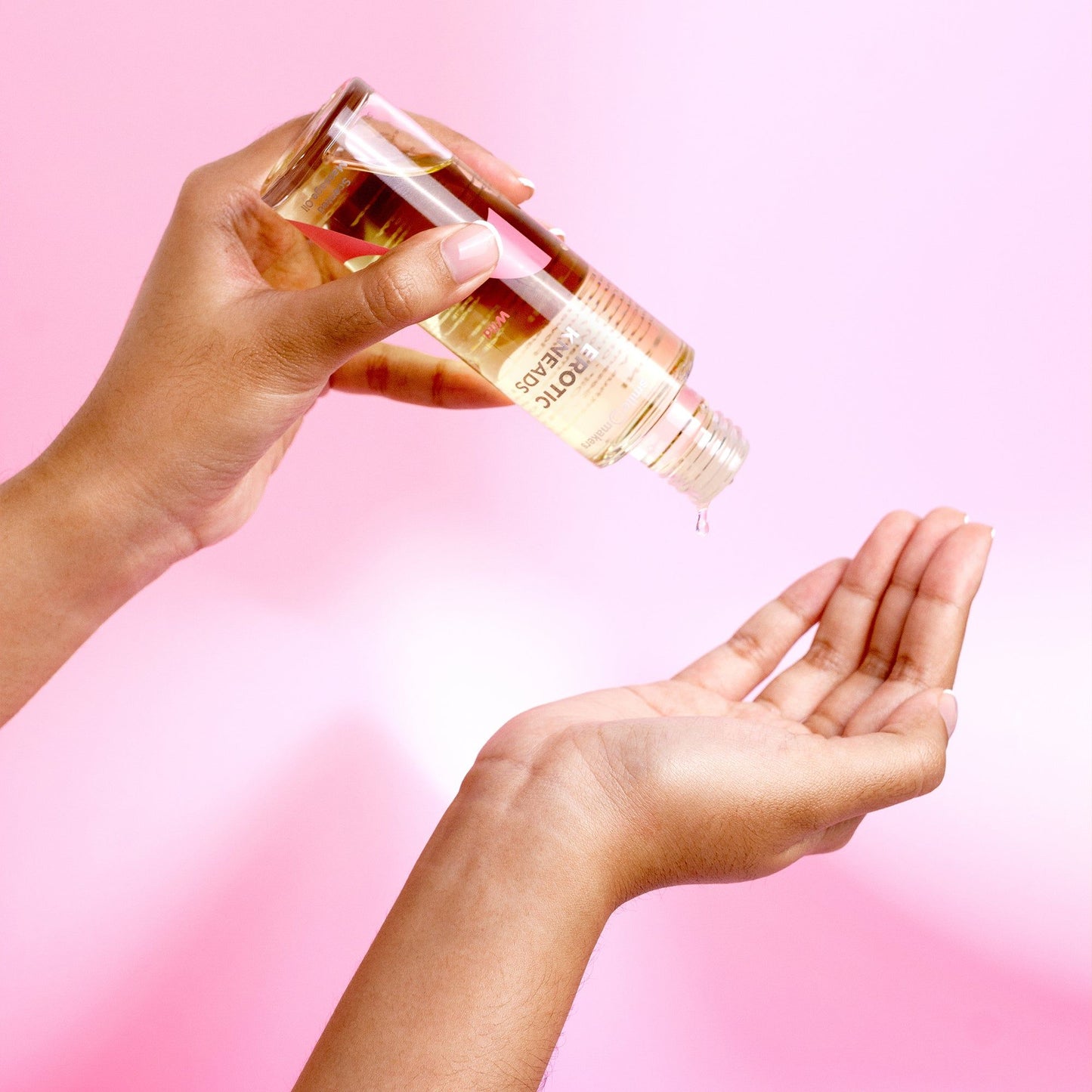 A person pours nourishing oil from a glass bottle into their open palm against a pink background. The glass bottle is transparent with a cylindrical shape and features a screw cap dispenser. The person’s hand is partially visible, holding the bottle upright above the other open palm. The background is solid light pink with no distinct texture or additional elements. Label text details are not visible in this image.