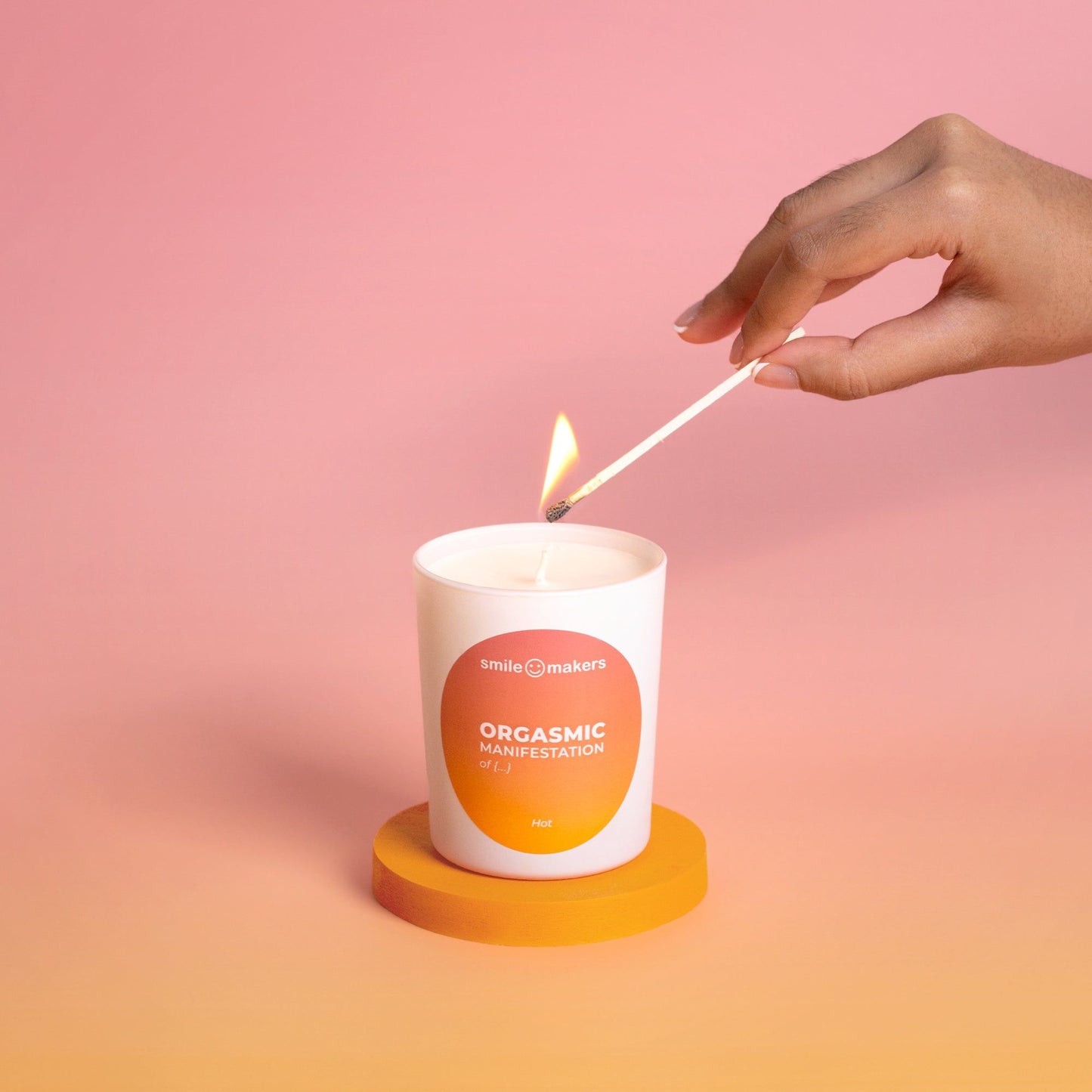 A hand uses a lighter to ignite a white cylindrical candle with the label “Orgasmic Manifestation by Smile Makers” in sans-serif font. The candle sits on a round orange coaster. The background has a bright pink and orange gradient. The image is taken as a close-up product shot.