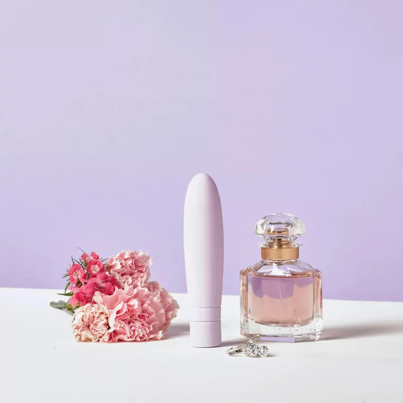 A white vibrator, a pink perfume bottle, a diamond ring, and pink flowers are artfully arranged on a white surface with a purple background. The vibrator is smooth with rounded edges and no visible branding. The perfume bottle is cube-shaped glass with a matte silver cap. The ring has a large central clear stone in a metal band. Flowers have full pink petals and green stems. No people or hands are present. No legible label text is visible. Shot is overhead top-down view with soft shadows.