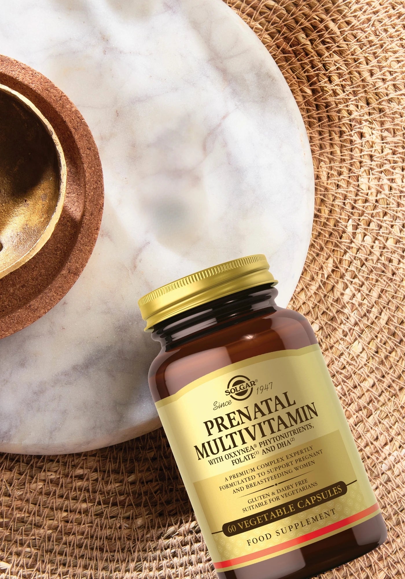Solgar Prenatal Multivitamin: pullo lepää kudotulla matolla marmori- ja korkkikulhon vieressä; pullo on ruskea kultaisella mattapintaisella etiketillä, mustalla ja punaisella versaaliserif-kirjoituksella, tekstissä lukee "Solgar" ja "Prenatal Multivitamin"; pakkaus mainitsee 60 kasvikapselia; ei ihmisiä tai käsiä näkyvissä; laaja kuvakulma otettu ylhäältä hieman viistosti.