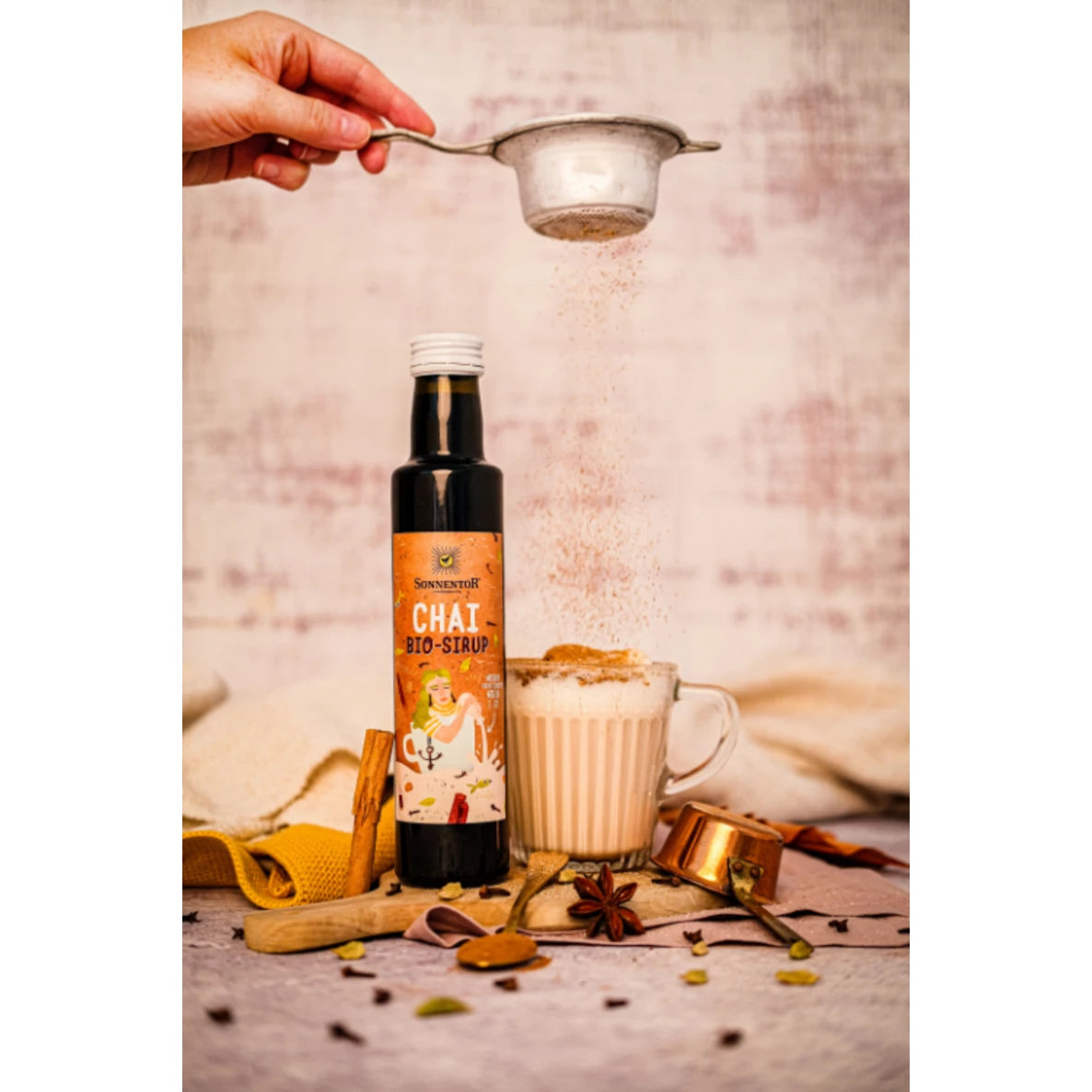 A hand sprinkles powder through a fine metal sieve over a clear glass mug filled with brown chai. Next to the mug is an amber glass bottle with a white label reading “Chai Bio-Sirup” in bold black sans-serif font. Whole spices, a stainless steel measuring cup, and a metal spoon rest on a light-colored tabletop. The shot is set indoors with even lighting. A hand sprinkles powder through a sieve over a glass mug of chai beside a bottle labeled Chai Bio-Sirup. Fragrant spices, a measuring cup, and a spoon are