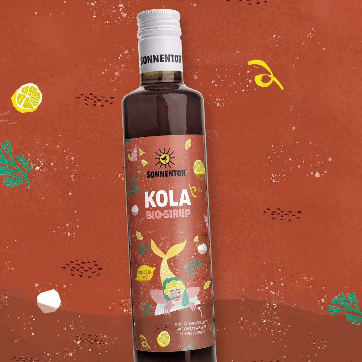 Pullo Sonnentor Kola Bio-Sirup -siirappia valkoisella muovisella kierrekorkilla seisoo mattaa punertavaa taustaa vasten, jossa on sarjakuvamaisia kuvituksia keltaisista sitruunaviipaleista, vihreistä lehdistä ja sinisistä kaloista. Pullo on lieriömäinen. Etiketissä on leikkisää grafiikkaa ja yksinkertaista pääteviivatonta tekstiä, joka kertoo kofeiinittomasta luomusiirapista.