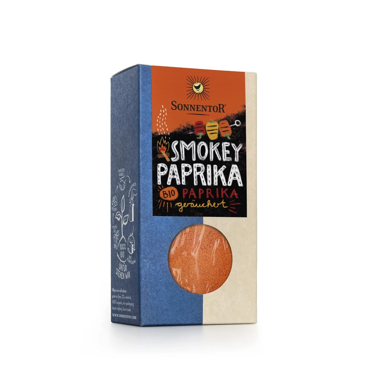Laatikko Sonnentor Smokey Paprika -jauhetta, jossa on värikästä brändäystä, pieni pyöreä ikkuna näyttämässä rikas paprika sisällä ja eloisia ruoka- ja keittiövälinekuvituksia. Pakkaus on suorakulmainen pahvi mattapinnalla. Etiketissä on tuotenimi lihavoiduin kirjaimin ja Sonnentor-logo yläosassa. Tausta on valkoinen ilman lisäesineitä tai ihmisiä.