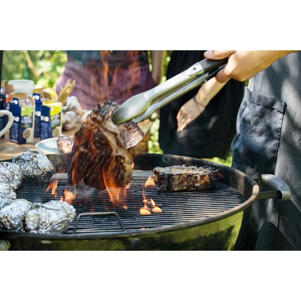 Henkilö käyttää pihtejä pihviä grillatessaan avotulella grillissä, lähellä näkyy foliopakattuja perunoita ja maukkaita kastikkeita. Grilli on musta metallia lankaritilällä. Pihvi on paksuksi leikattua, ruskeaa grillijäljin. Perunat on käärittynä kiiltävään alumiinifolioon. Kastikepullot ovat muovia, puristettavin korkein ja luettavin etikettein ulospäin. Tausta on ulkona.