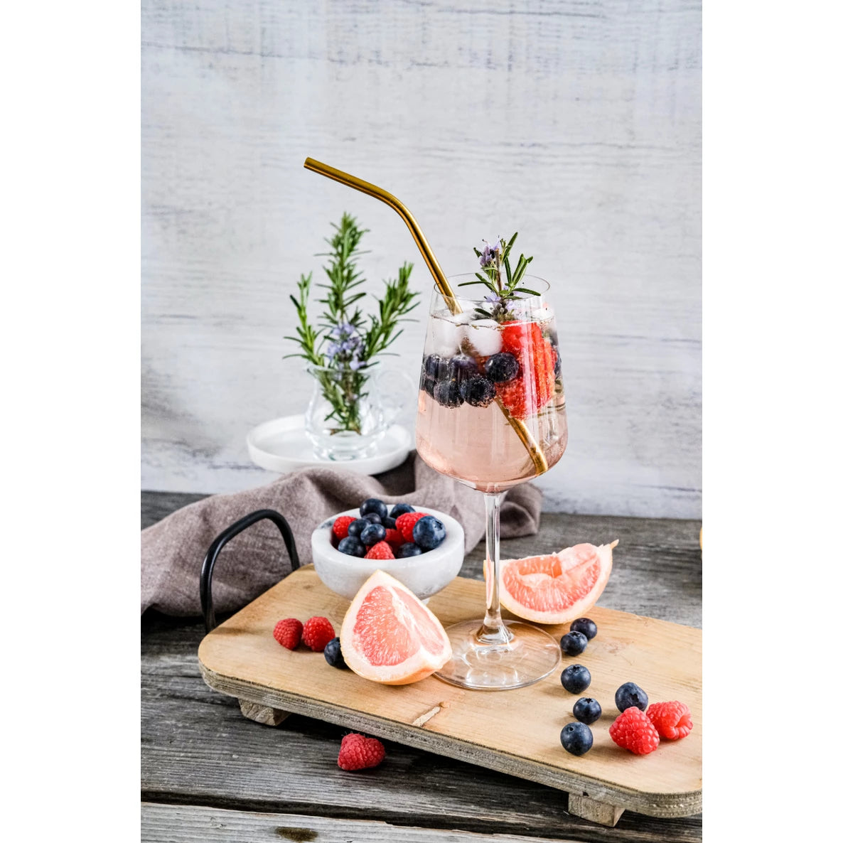 Lasi vaaleanpunaista cocktailia marjoin, jääpaloin ja rosmariininoksalla seisoo puulaudalla greippiviipaleiden, marjojen ja kultaisen pillin kanssa. Lasi on kirkas, sileäpintainen ja pyöreämuotoinen. Lauta on luonnonpuuta näkyvin syin. Lähellä on siroteltuja marjoja ja sitrushedelmäviipaleita. Taustalla kulhossa on tuoreita marjoja; lisää rosmariininoksia on nähtävissä. Etikettejä tai tekstiä ei näy missään kohtauksessa. Ihmisiä tai käsiä ei näy kuvassa.