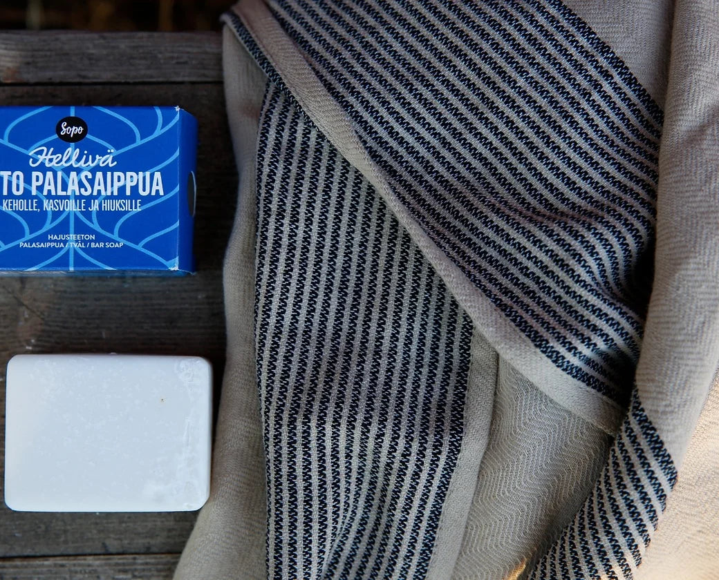 Sopo Hellivä Aito Palasaippua Hajusteeton; sininen suorakaiteen muotoinen saippualaatikko mattapinnalla, valkoinen neliömäinen kiinteä saippuapala sileine reunoineen sekä beige puuvillapyyhe mustin suuntaisviivoin, aseteltu vierekkäin keskikarkealle puupinnalle; ei näkyvää typografiaa tai etikettitekstiä; ei ihmisiä eikä käsiä näkyvissä; siniset, valkoiset ja beiget tuotteet järjestetty siististi puisen alustan päälle.