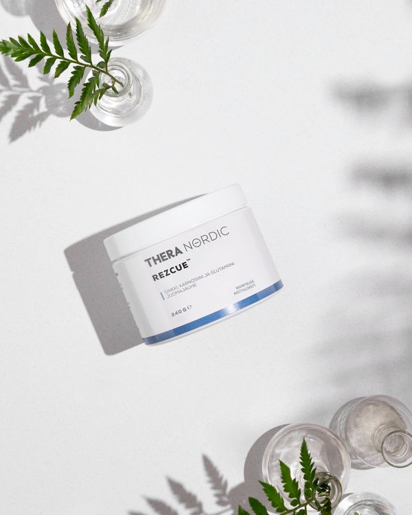 THERA Nordic Rezcue; valkoinen, mattapintainen sylinterinmuotoinen purkki, jossa lukee isoin kirjaimin lihavalla groteskilla "THERA NORDIC RESCUE", seisoo tasaisella valkoisella pinnalla; lähellä eri korkuisia läpinäkyviä lasimaljakoita; maljakoissa tuoreita vihreitä lehtiä, joiden varjot lankeavat pinnalle luonnonvalossa; kuvassa ei näy ihmisiä tai käsiä.