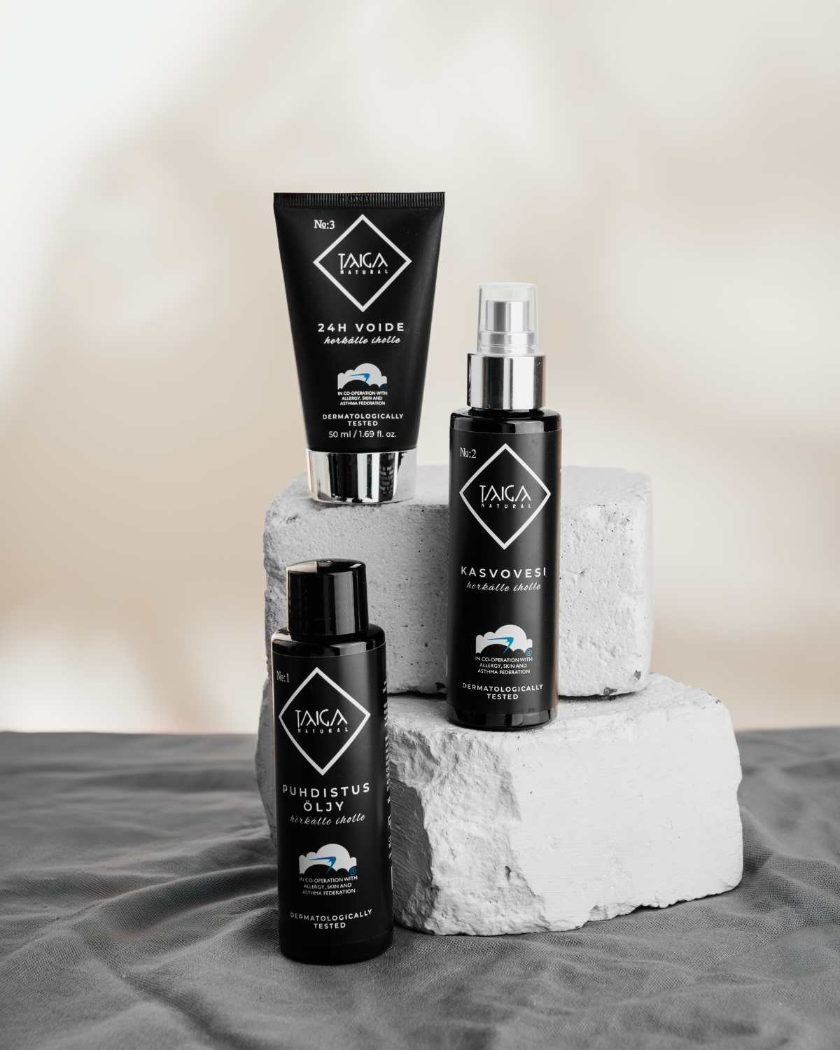Taiga Cosmetics Kasvovesi; kolme mustaa Taiga-brändin ihonhoitopulloa mattapinnalla aseteltuna yhtenäisille valkoisille kivilohkoille; astiat ovat sylinterin- ja suorakulmaisen muotoisia pumppukorkein tai kansin; etiketit ovat valkoisia ja sinisiä selkeällä yksinkertaisella groteskikirjasimella, osittain näkyvä brändinimi "Taiga"; tuotteet seisovat eri korkeuksilla; tausta neutraali ja epätarkka; ei ihmisiä tai käsiä näkyvissä, studioasetelma.