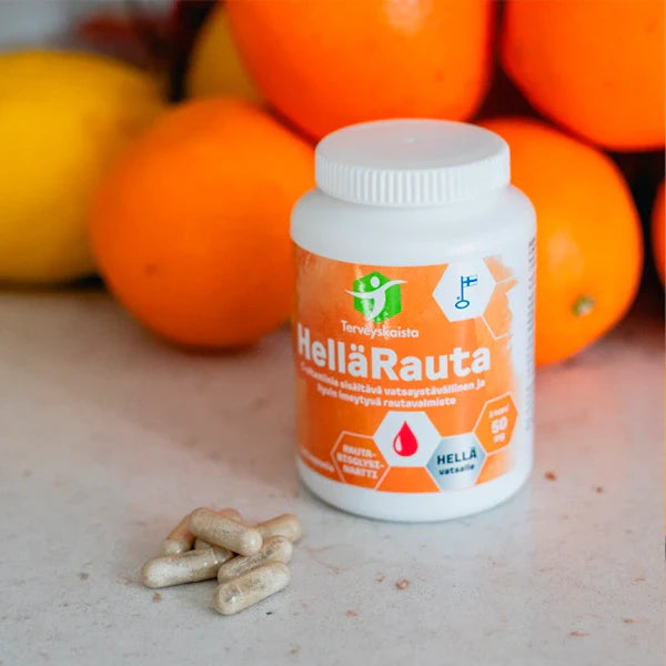 Terveyskaista HelläRauta 50 mg: sylinterimäinen muovipullo mattaetiketillä tekstillä "HellaRauta" seisoo sileällä tasolla; muutama tumma rautakapseli hajallaan pullon vieressä; taustalla epätarkkoina appelsiineja ja sitruunoita; ympäristö on yksinkertainen sisätila tasaisella valaistuksella; ei ihmisiä tai käsiä näkyvissä.