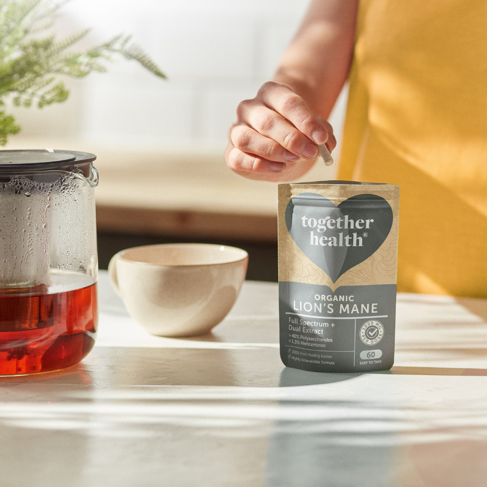 Together Health Lions Mane; henkilö valmistaa teetä kirkkaan lasisen teekannun ja valkoisen kupin vieressä puupöydällä vahvassa luonnonvalossa; mattapintainen seisova pussi tekstillä ”Together Health Organic Lions Mane” pystyssä lihavoidulla antiikvatekstillä ja niukoin graafisin elementein; ei irtokapseleita tai tabletteja näkyvissä; tausta epätarkka kodinomainen tila.