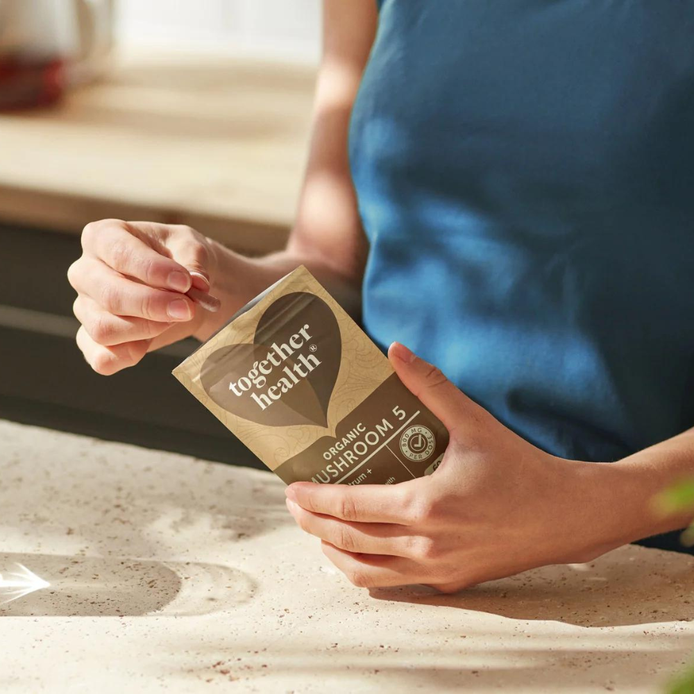 Together Health Mushroom 5; sinisessä paidassa oleva henkilö avaa mattapintaisen ruskean taipuisan pussin tekstillä ”Together Health Organic Mushroom 15” lihavoidulla valkoisella ja vihreällä groteskitekstillä, vaalealla keittiön työtasolla; pakkaus suorakaiteen muotoinen uudelleensuljettavalla reunalla, ei näkyvää ikkunaa tai grafiikkaa; henkilö avaa ruskean pussin korostaen luonnonmukaisia ainesosia vaalealla työtasolla.