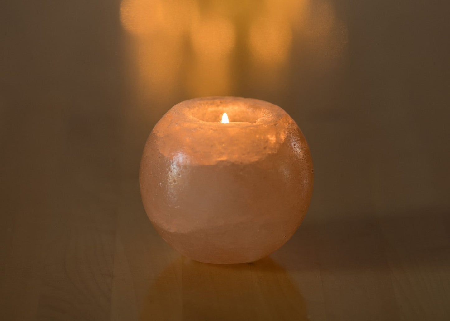 A lit candle burns inside a round, pink Himalayan salt holder, casting a soft, warm glow on a smooth brown surface. The holder is solid and has a matte finish. There is no visible label or packaging. The background and setting are minimal with only the surface visible, and there are no people or hands in the shot.
