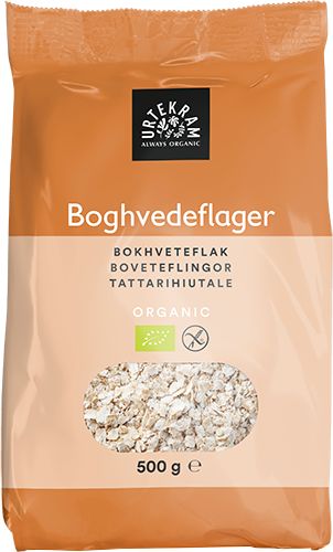 A 500g bag of Urtekram organic buckwheat flakes with a transparent window displaying the nutritious flakes inside. Rectangular, upright pouch format; packaging is matte orange with a clear section on the front. Sans-serif text in white and dark colors shows product name and multiple languages. No people, plain background, studio product shot.