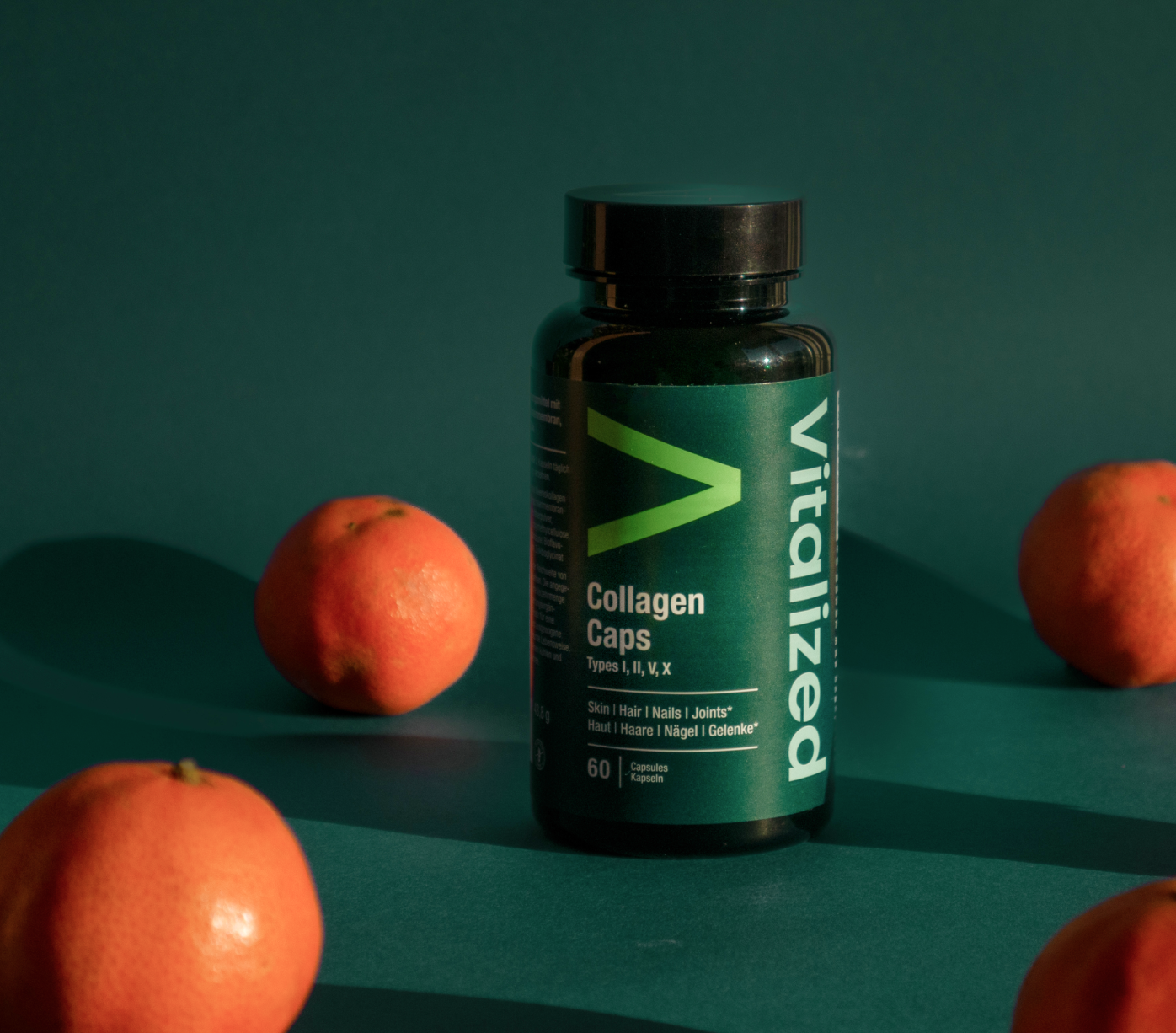 Vitalized Collagen Caps; lieriömäinen muovipullo tekstillä Vitalized Collagen Caps seisoo pystyssä sileällä, yhtenäisellä vihreällä pinnalla; neljä kuorimatonta mandariinia aseteltuna pullon ympärille; pullo mattavalkoinen tummalla kannella; etiketissä groteskitekstiä, joka kertoo hyödyistä iholle, hiuksille, kynsille ja nivelille; ei ihmisiä tai käsiä; tuotekuva kaikkien esineiden näkyessä yksinkertaista taustaa vasten.