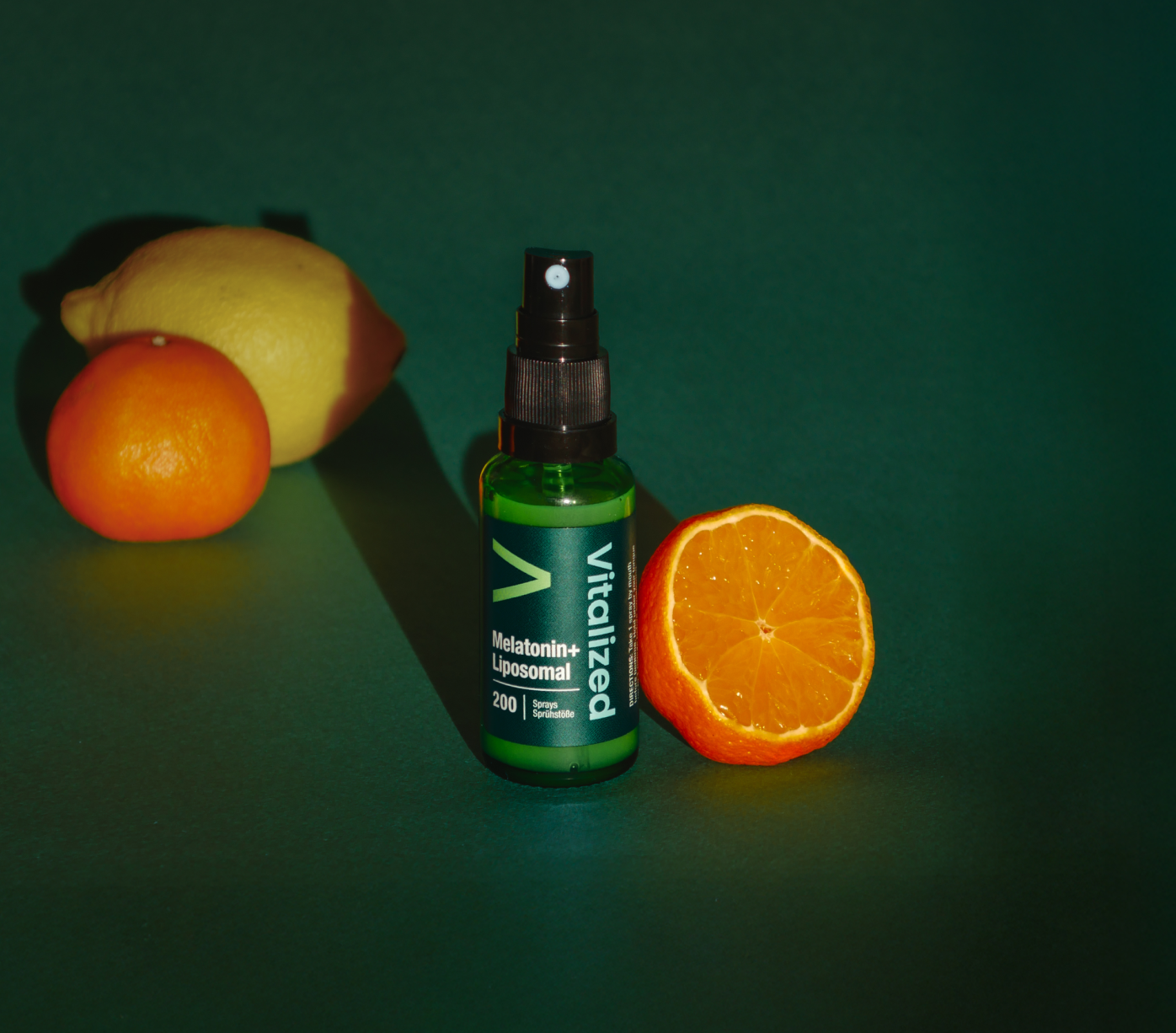 Vitalized Melatonin+ Liposomal; vihreä pullo tekstillä ”vitalized Melatonin+ Liposomal” seisoo viipaloidun appelsiinin, kokonaisen appelsiinin ja sitruunan vieressä tummanvihreällä taustalla; pullo sylinterin muotoinen sileäpintainen valkoisella groteskitekstillä etiketissä; ei ihmisiä kuvassa; tuote ja hedelmät aseteltuna pystyyn studioasetelmassa.