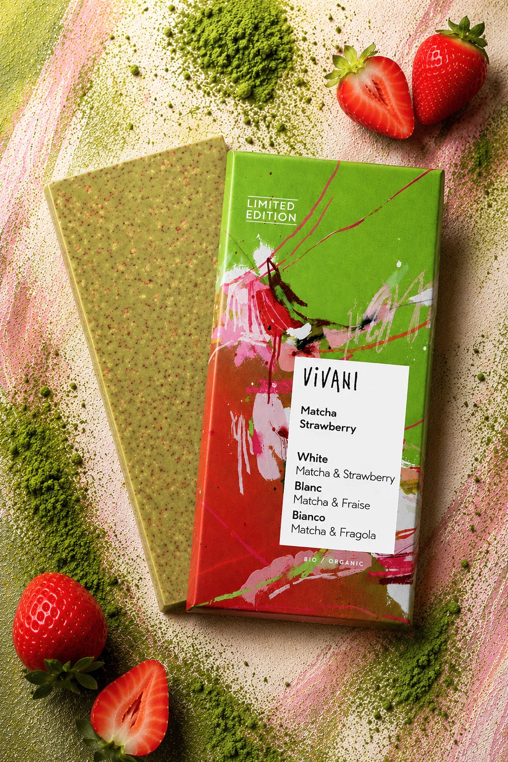 Vivani Matcha Strawberry -suklaapatukka, osittain avattu, lepää vaaleanpunaisella ja vihreällä jauhemaisella alustalla viipaloitujen ja kokonaisten mansikoiden ja eloisan matchajauheen ympäröimänä. Suklaapatukka on suorakulmainen mattapinnalla, väriltään vaaleanvihreä. Pakkauksessa on yksinkertainen mustavalkoinen typografia. Ihmisiä tai käsiä ei näy, kuva on otettu ylhäältä.