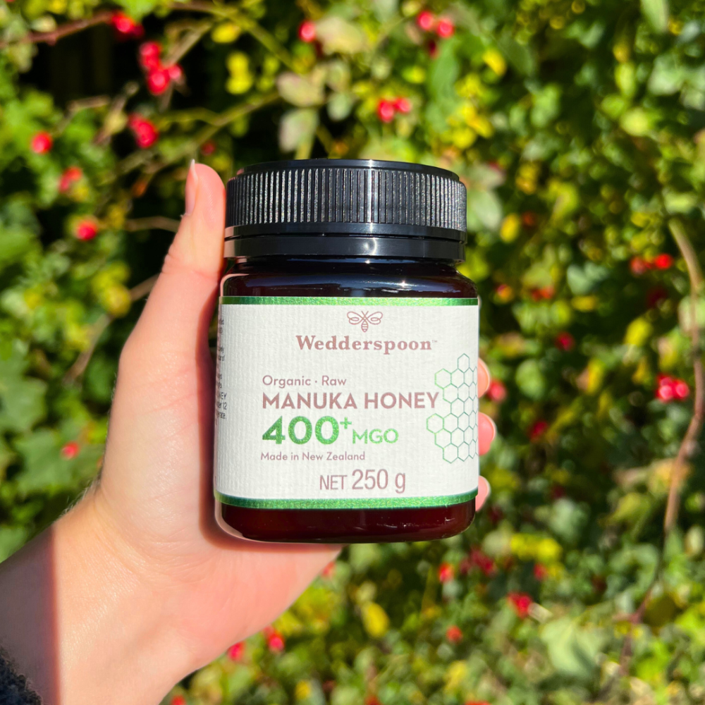 Wedderspoon Luomu Manuka-hunaja 400+ MGO; käsi pitelee muovipurkkia Wedderspoon Organic Raw Manuka Honey 400+ MGO, mustalla kierrekannella ja vaaleanpunaisella mattaetiketillä valko- ja oranssisävyisin groteskitekstein; pääetiketissä tuotenimi selkeästi esillä; taustalla epätarkkaa vihreää kasvillisuutta ja punaisia marjoja; lähikuva.