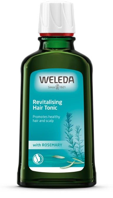 A green glass bottle contains Weleda Revitalising Hair Tonic with Rosemary. The bottle has a rectangular shape with rounded edges and a white cap. The front label is blue, matte, and displays a detailed rosemary illustration with sans-serif white text. The label is centered on the front face. Background is plain white; no people or hands are present in the image. A green bottle of Weleda Revitalising Hair Tonic with Rosemary is designed to promote healthy hair and scalp. The front features a blue label