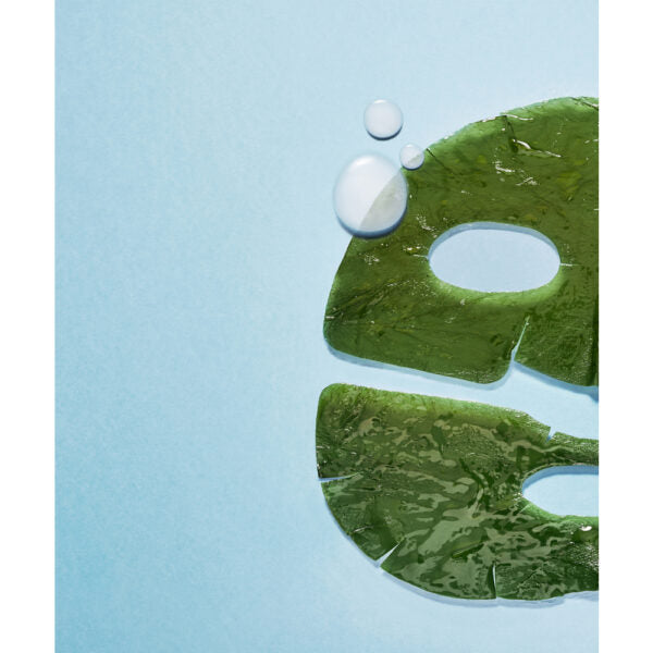 Whamisa Algae Kelp Mask; vihreä kangasnaamio vesipisaroineen vaaleansinisellä taustalla, kuvattu rajattuna kulmasta, jossa näkyvissä silmä- ja suuaukot kosteutusta ja virkistystä varten; naamion pinta sileä ja kostea, aseteltu tasaisesti; ei pakkausta, tekstiä tai ihmisiä näkyvissä; tausta yksivärinen ja tasaisesti valaistu; kuva läheltä ja ylhäältä kuvattuna.