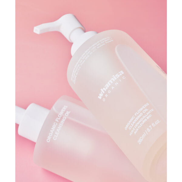 Whamisa Organic Flowers Cleansing Oil; kaksi Whamisa Organic Flower Cleansing Oil -pulloa valkoisin pumpuin vaaleanpunaisella taustalla, toinen pystyssä ja toinen kyljellään; pullot läpikuultavia huurretuin, mattapintaisin lieriömäisin muodoin; pakkauksessa valkoista kukka-aiheista tekstiä sekä englanniksi että korealaisin kirjaimin; ei ihmisiä tai käsiä kuvassa.