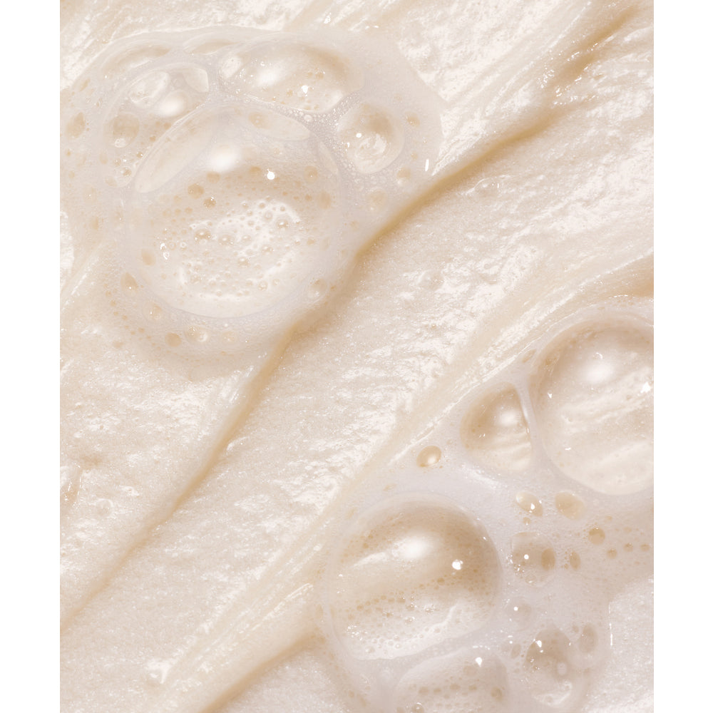 Whamisa Organic Flowers Foam Cleansing Cream; lähikuva kermaisesta, beigenvärisestä vaahtoavasta tuotteesta, jonka pinnalla kuplia; sileä ja kiiltävä tekstuuri, siellä täällä isompia ja pienempiä kuplia; ei näkyvää pakkausta, tekstiä tai taustaa; ei ihmisiä tai käsiä; kuva keskittyy tiiviisti nesteen pintatekstuuriin.