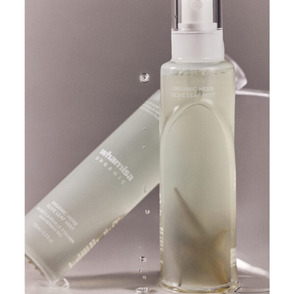 Whamisa Organic Flowers Olive Leaf Mist; kaksi Whamisa Organic Herb Olive Leaf Mist -pulloa, toinen pystyssä ja toinen vaakatasossa; pakkaus koostuu läpinäkyvistä lieriömäisistä pulloista, joissa vihertäväsävyistä nestettä, ja mattavalkoisista korkeista; ainakin yhdessä pullossa näkyvissä yksinkertaista groteskibrändäystä; vesipisaroita esineiden ympärillä tasaisella taustalla; ei ihmisiä tai käsiä kuvassa.