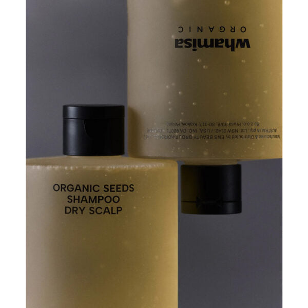 Whamisa Organic Seeds Shampoo Dry Scalp; kaksi beigeä Whamisa Organic Seeds Shampoo -pulloa kuivalle päänahalle, rikastettu luonnonaineksin, esillä lähikuvassa; pullot mattamuovia mustin läppäkorkein; kummassakin niukkaa etusivun tekstiä yksinkertaisella groteskityylillä; toinen pullo pystyssä, toinen ylösalaisin tasaisella taustalla.