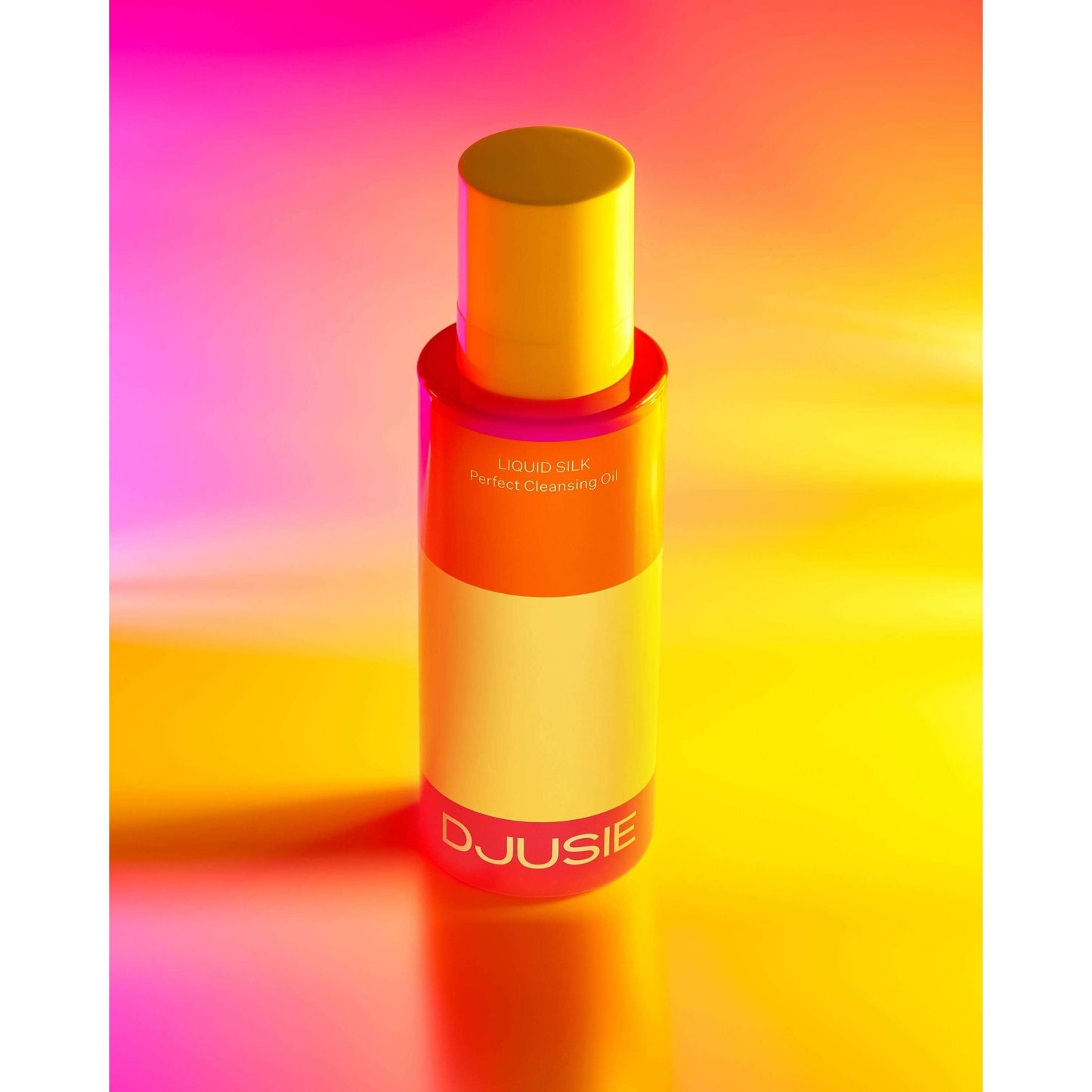 A bottle of DJUSIE Liquid Silk Perfect Cleansing Oil stands upright on a vibrant yellow and pink gradient background, casting a soft, luminous shadow. The bottle is cylindrical with a pump top and transparent body showing pale yellow liquid inside. The label displays product name in bold sans-serif text. No people or hands are visible in the image.