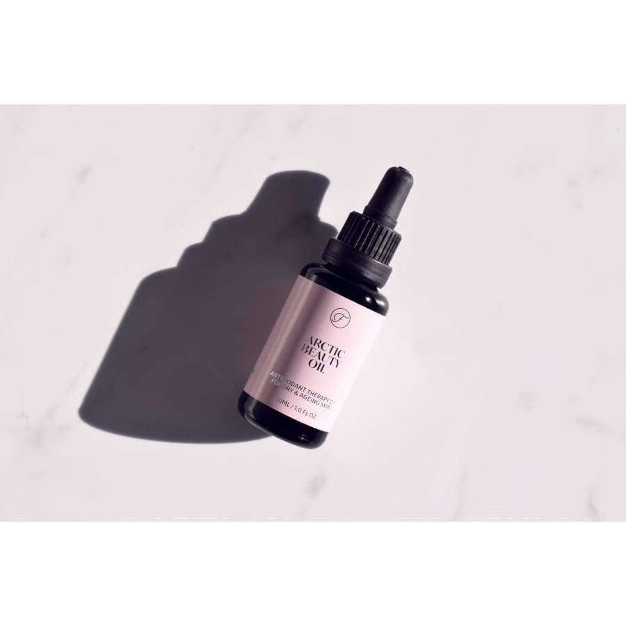 FLOW Cosmetics Arctic Beauty Oil -tiputinpullo tummaa lasia, lepää vaalealla pinnalla pehmeän varjon kanssa; lieriömäinen, musta kierrekorkki ja kiinteä tiputin; etiketissä vähän, selkeää tekstiä; tausta tekstuuriton valkoinen; kuvassa vain tuote, ei rekvisiittaa tai ihmisiä.