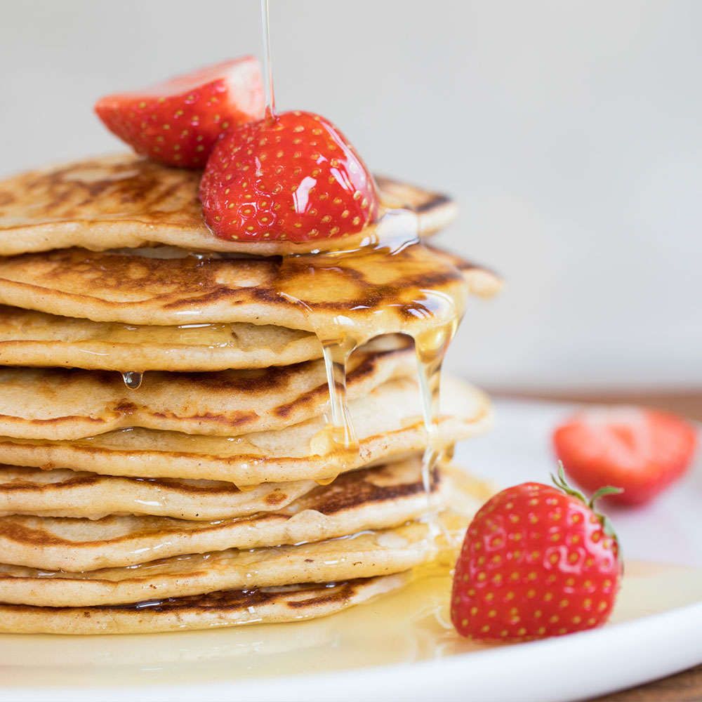 A stack of pancakes topped with fresh strawberries and syrup being poured over them, with extra strawberries scattered on the plate. Pancakes are round and golden brown with a matte finish; syrup is clear amber. No packaging or typography is visible. The background is a plain tabletop surface, shot from slightly above. No people or hands shown.