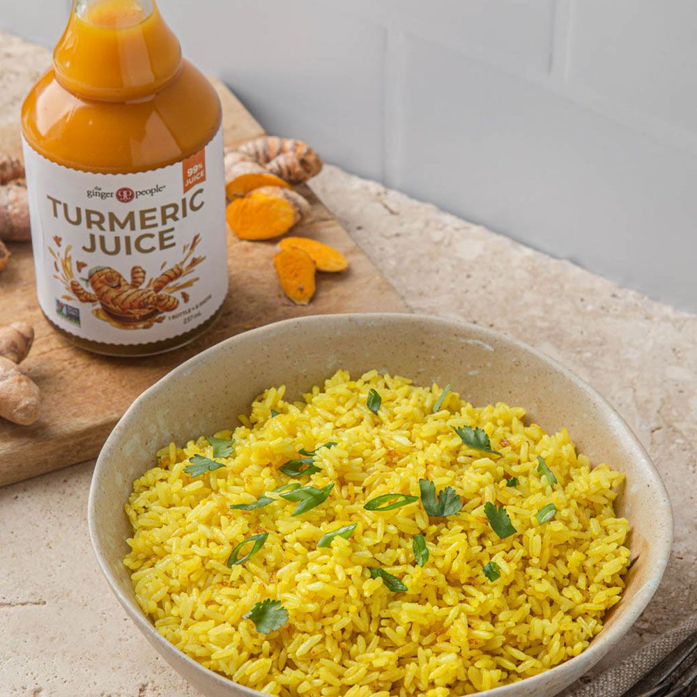 A bowl of vibrant yellow turmeric rice with green herb garnish is placed beside a clear glass bottle filled with golden-yellow liquid, labeled as turmeric juice in simple, sans serif font. Several slices of raw orange turmeric root are on the wooden board. The setting is a tabletop scene without people shown. A bowl of vibrant yellow turmeric rice garnished with herbs sits next to a bottle of golden turmeric juice and slices of fresh turmeric root on a wooden board.