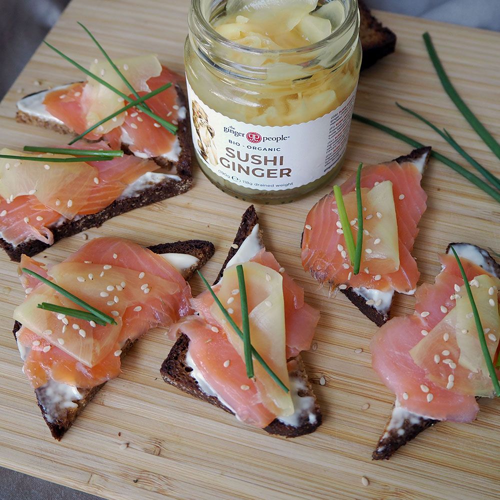 Open-faced sandwiches with smoked salmon, sushi ginger, chives, and sesame seeds on brown bread slices surround an open jar of sushi ginger on a wooden board. The jar is glass with a metal lid, partially filled with pink sliced ginger; its label includes legible product information in simple sans-serif type. The background is plain wood. No people or hands are visible in the shot.