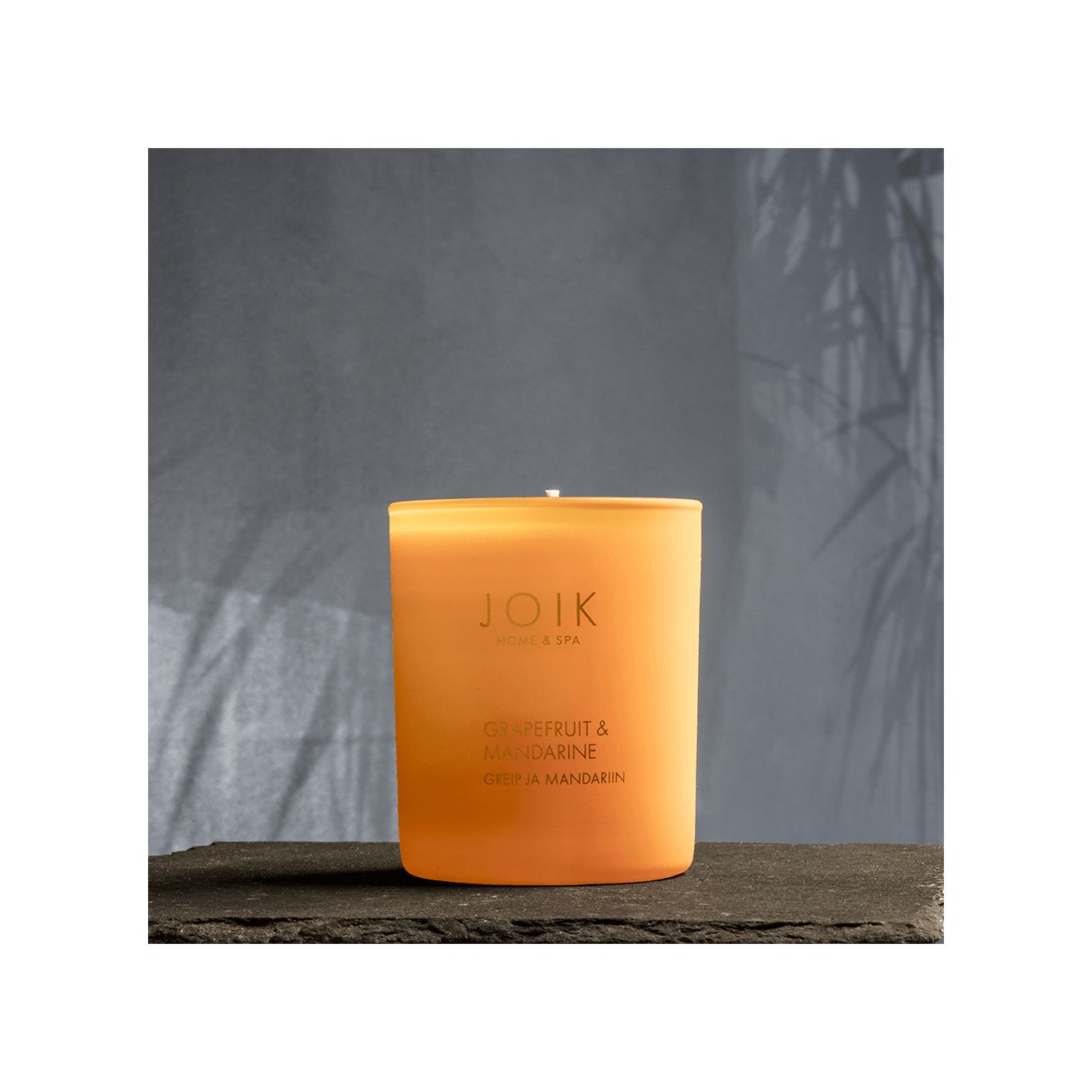 An orange cylindrical candle in a matte glass container shows the text “JOIK Home & Spa” and “Grapefruit & Mandarin” on a white rectangular label with simple sans-serif font. The candle sits on a smooth, dark surface. The background is gray with diffuse shadows of foliage. No people or hands are visible in the image.