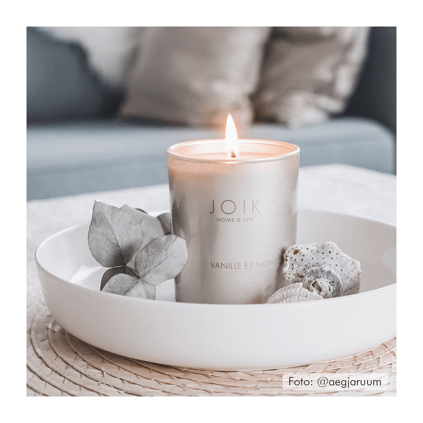 A lit JOIK Home & Spa candle in a cylindrical glass container with a matte white label displaying the product name in sans-serif type sits centered in a glossy white shallow dish. The dish is surrounded by decorative green leaves and a smooth, oval gray stone. The entire arrangement rests on a tan woven mat with visible texture. Background features an out-of-focus upholstered sofa in light color tones. No people or hands are present in the image.
