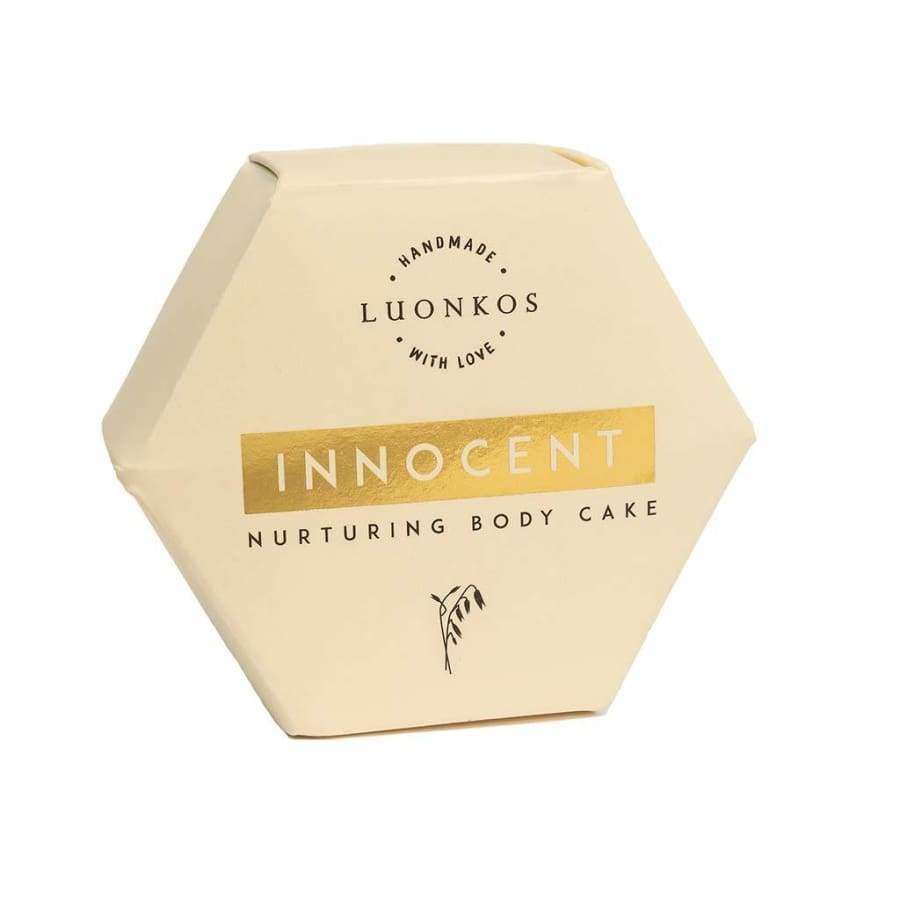 Luonkos Innocent Nurturing Body Cake; kuusikulmainen kermanvärinen pakkaus kultaisin korostuksin ja pelkistetyllä kaurakuvituksella hoitavan vaikutuksen viestimiseksi; pakkaus mattapintainen, terävin reunoin, painetut elementit mustana ja kultaisena; groteskityyppiä; tuote kuvattu yksin vaalealla taustalla, ei ihmisiä tai käsiä.