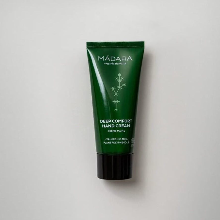 A green plastic tube of MÁDARA Deep Comfort Hand Cream with a black screw-on cap, rectangular shape, and smooth matte surface. White sans-serif label text includes brand name and product type. No additional graphics or icons visible. The tube is upright on a plain white background. No people or hands present. A green tube of MÁDARA Deep Comfort Hand Cream with a black cap, featuring nourishing botanical extracts, displayed on a plain white background.
