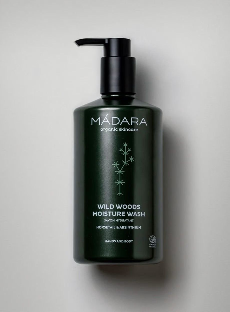 A dark green plastic pump bottle with a matte finish, cylindrical shape, and flat base. The label shows Mádara Wild Woods Moisture Wash with organic skincare, mentions horsetail and absinthium, and states it is suitable for hands and body. The typography is modern sans serif in white. Bottle stands on a smooth light gray background; no people or hands visible.