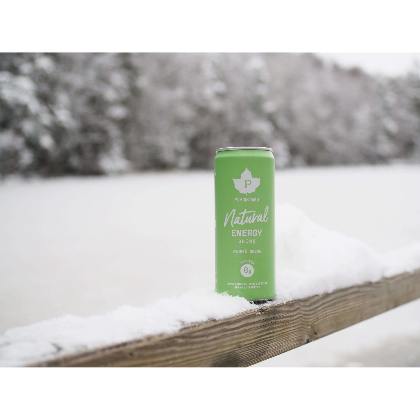 Puhdistamo Natural Energy Drink, Vihreä Omena; vihreä alumiinitölkki mattapinnalla, lieriömäinen, luettavissa lihavoitua groteskitekstiä brändinimestä; tölkki asetettu pystyyn lumisen puukaiteen päälle, taustalla epätarkka metsämaisema; ei ihmisiä tai käsiä kuvassa.