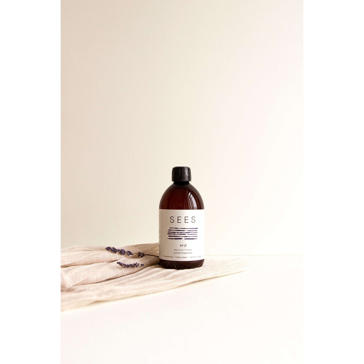 Amber bottle labeled SEES with a black cap sits on a beige cloth with lavender sprigs, against a plain light background. The bottle is cylindrical and made of amber glass with a matte finish. The cap is opaque black plastic. “SEES” is printed in bold, uppercase letters centered on the label. No people or hands are present in the image.