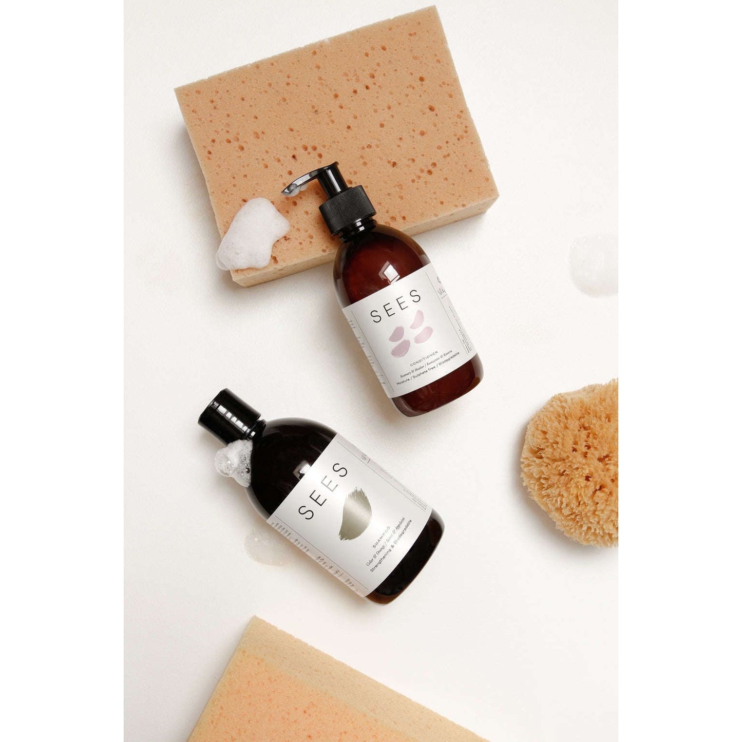 Two brown bottles of SEES skincare products rest on a white surface with beige bath sponges. The first bottle has a pump dispenser with soft foam visible at the opening; the second has a flip cap and small bubbles near the top. Both bottles have simple sans-serif SEES branding printed in white. Water droplets are present on the surface nearby, no people or hands appear in the image.