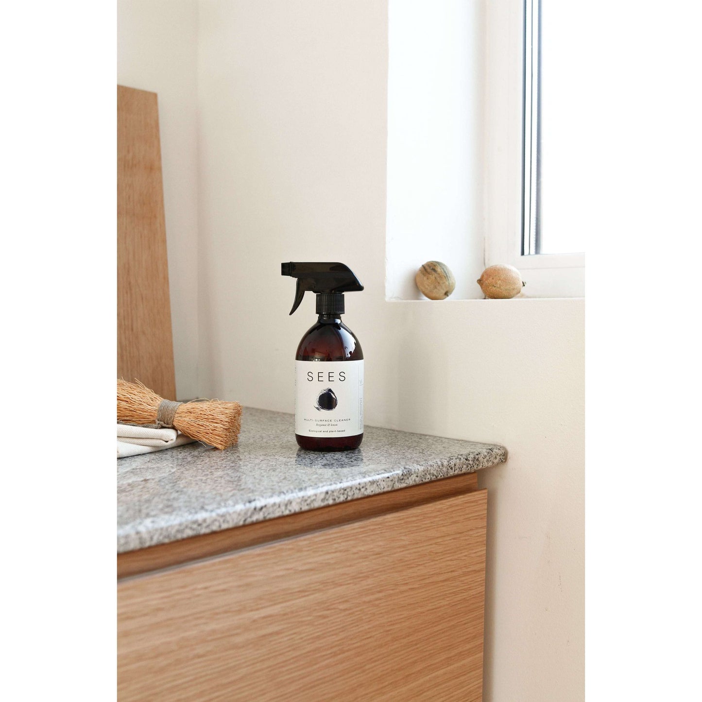 A transparent plastic spray bottle labeled SEES with black block letters stands on a polished granite countertop. A small broom with natural bristles and wood handle lies beside it. The background includes light-colored wood surfaces, a window ledge with three smooth round stones, and diffused natural light. There are no people or hands present in the image.