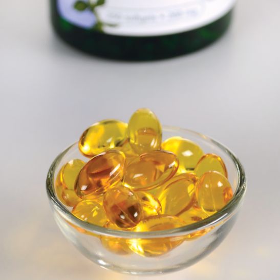 A small glass bowl filled with golden-yellow vitamin softgel capsules sits on a light surface; a blurred bottle is visible in the background. The capsules are oval and translucent with a glossy finish. The bottle in the background has no clearly readable label text due to blurring. No people or hands are present. The image is shot from above at an angle.