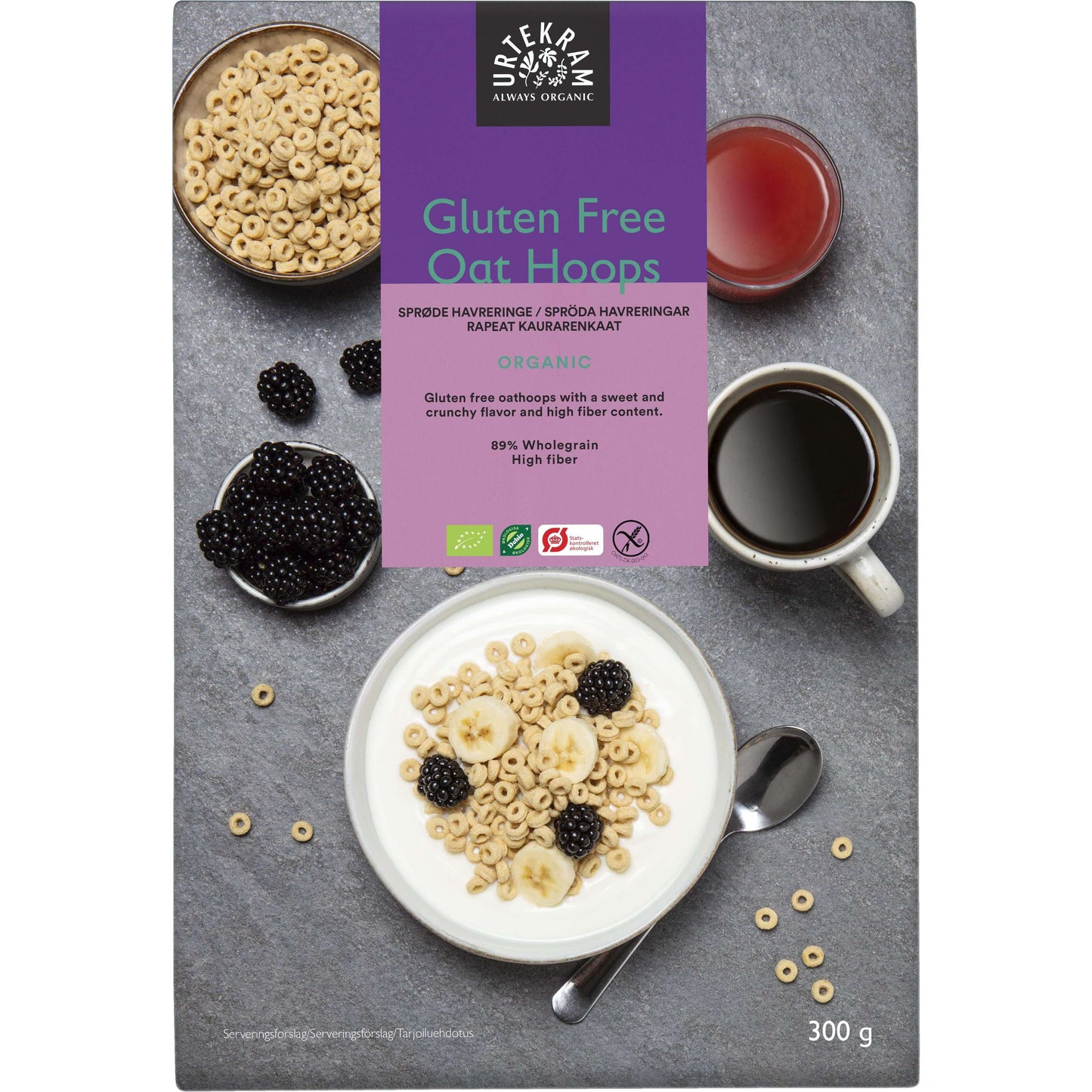 Rasia Urtekram Gluten Free Oat Hoops -mysliä, kuvituksena kulhollinen mysliä banaaniviipaleiden ja karhunvatukoiden kera, kuppi teetä, purkki hilloa ja kulhollinen luomukarhunvatukoita. Rasia painaa 300 g. Myslirasia on suorakulmainen mattapinnalla, pääosin vihreä, luettava tuote- ja brändinimi lihavoidulla pääteviivattomalla fontilla. Esineet on aseteltu vaalealle tasaiselle alustalle pöytäasetelmana, ei ihmisiä tai käsiä näkyvissä.