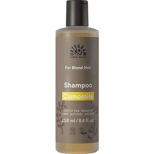 Urtekram Kamomilla Shampoo; pullo tekstillä "Urtekram Nordic Beauty Chamomile Shampoo" vaaleille hiuksille, 250 ml, lieriömäinen muoviastia mattaharmaalla ja keltaisella etiketillä; etiketissä tuotetietoja, mukaan lukien luomu, vegaanisertifiointi ja 100-prosenttisen luonnollinen alkuperä modernilla groteskitypografialla; tausta yksinkertainen, ei ihmisiä tai käsiä näkyvissä.