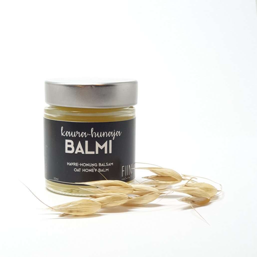 Fiini Naturally Kaura-Hunaja Voide; purkki tekstillä "Oat Honey Balm", kansi heijastavan hopeinen; purkki sylinterinmuotoinen ja kirkas tai huurteinen; etiketti yksinkertaisella lihavoidulla groteskikirjasimella; vierellä kaurantähkiä ja hunajapisaroita valkoisella alustalla; tasaisesti valaistu studiokuva ilman ihmisiä tai käsiä.